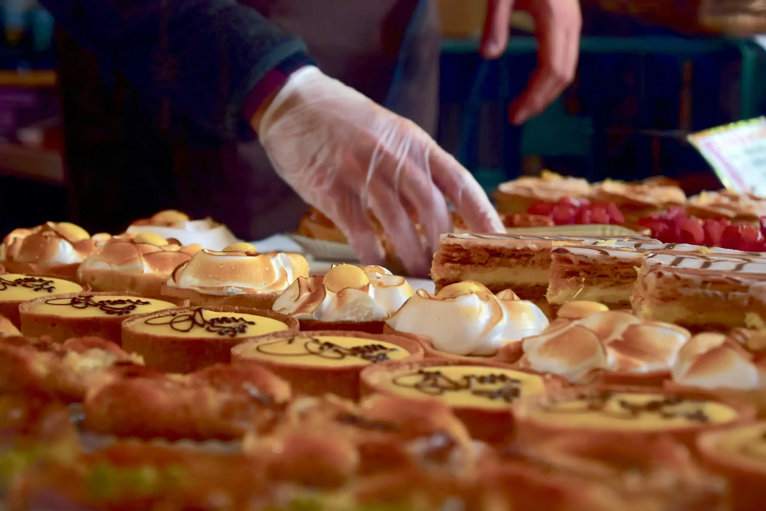 A pastry chef wearing gloves arranges an assortment of Delicious Breakfast Pastries, including lemon tarts, mille-feuille, and meringue-topped treats, at a bakery display.